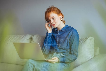 Téléchargez les photos : Jeune femme avec ordinateur portable et lunettes assis sur le canapé à la maison - en image libre de droit