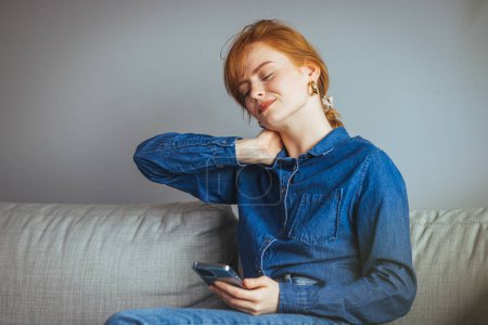 Téléchargez les photos : Frustré jeune femme souffrant de douleur au cou massages son cou tout en étant assis sur le canapé - en image libre de droit