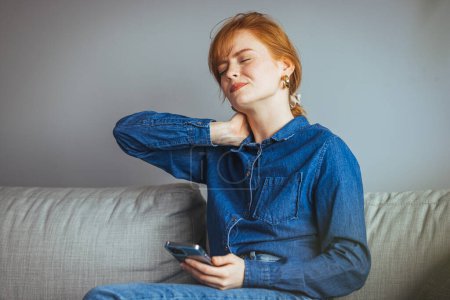 Téléchargez les photos : Frustré jeune femme souffrant de douleur au cou massages son cou tout en étant assis sur le canapé - en image libre de droit