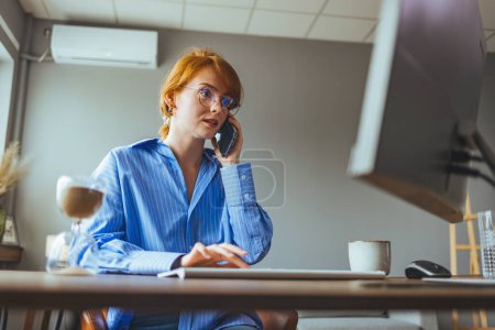 Foto de Hermosa joven empresaria que tiene una llamada de negocios en la oficina, su lugar de trabajo, anotando algo de información. - Imagen libre de derechos