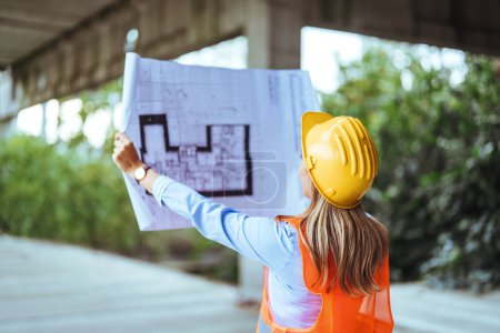 Photo for Back view of female construction worker in yellow safety helmet holding blueprint at the construction site - Royalty Free Image