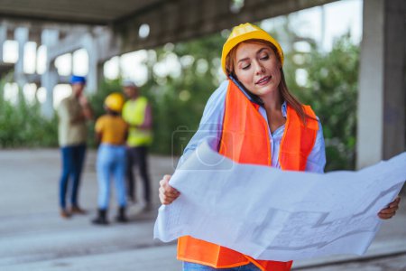 Photo for Caucasian female worker using smartphone and holding blueprint on construction site - Royalty Free Image