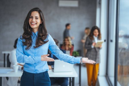 Photo for Happy young caucasian businesswoman showing welcome sign with colleagues on background in blurred office - Royalty Free Image
