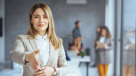 Téléchargez les photos : Heureuse jeune femme d'affaires caucasienne montrant signe de poignée de main avec des collègues sur fond dans le bureau flou - en image libre de droit