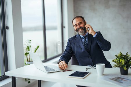Photo for Smiling caucasian businessman talking on smartphone while working at desk in office - Royalty Free Image