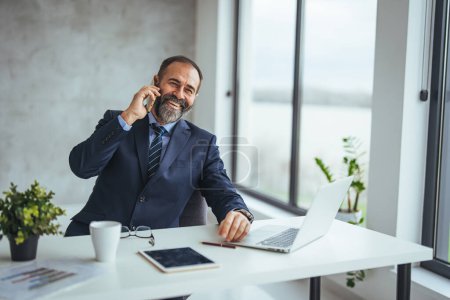 Photo for Smiling caucasian businessman talking on smartphone while working at desk in office - Royalty Free Image