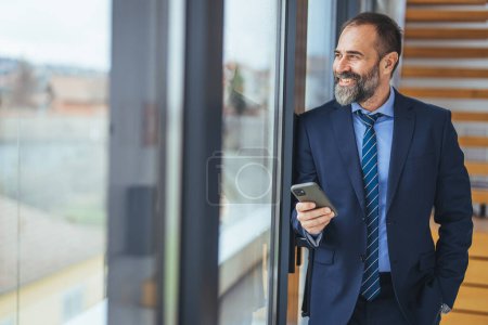 Foto de Hombre de negocios maduro sonriente sosteniendo un teléfono inteligente en una oficina. Empresario mirando a la cámara mientras está solo en un lugar de trabajo moderno. Hombre de negocios experimentado que se comunica con sus clientes. - Imagen libre de derechos