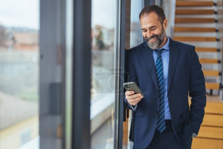 Téléchargez les photos : Homme d'affaires mature souriant tenant un smartphone dans un bureau. Homme d'affaires regardant la caméra tout en restant seul dans un lieu de travail moderne. Homme d'affaires expérimenté communiquant avec ses clients. - en image libre de droit