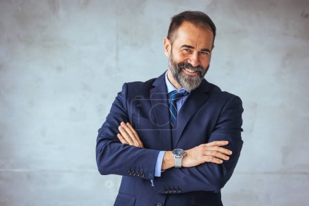Photo for Portrait of a happy mature business professional standing with his arms crossed in modern office. Smiling businessman at his workplace. Successful Mature man with folded arms standing over grey background. - Royalty Free Image