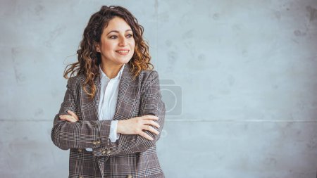 Photo for Portrait of middle aged businesswoman in modern office looking at camera. Confident business woman with arms crossed standing while leaning against glass wall. Proud brunette woman smiling in formalwear with copy space. - Royalty Free Image