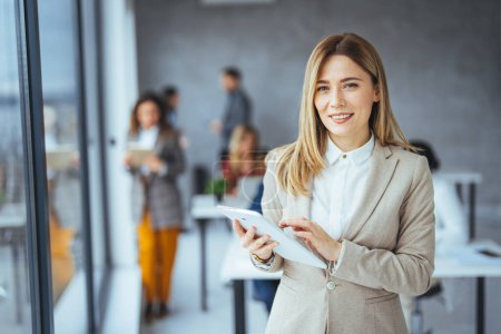 Photo for Young attractive female manager working on digital tablet while standing in modern office. Confident business expert. Attractive young smiling woman in smart casual wear holding digital tablet and looking at camera - Royalty Free Image