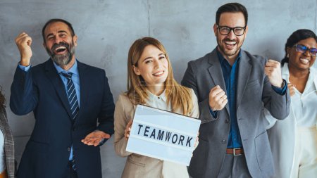 Photo for Portrait of multi ethnic successful business team looking at camera in the office and team leader holding sign ''teamwork'' in hands. Diverse creative team looking happy - Royalty Free Image