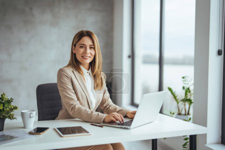 Photo for Successful smiling mature businesswoman using laptop and computer while doing some paperwork at the office. Young female professional at desk smiling to camera - Royalty Free Image