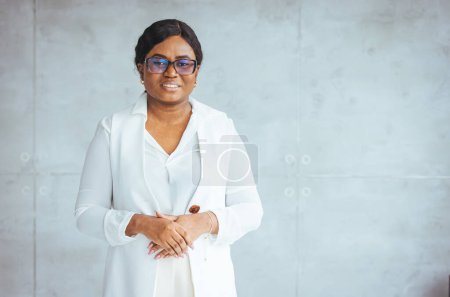 Foto de Retrato de una mujer de negocios afroamericana sonriente con gafas con estilo que mira a la cámara de pie en la oficina moderna. Concepto exitoso de negocio y carrera. Joven mujer de negocios en la oficina moderna - Imagen libre de derechos
