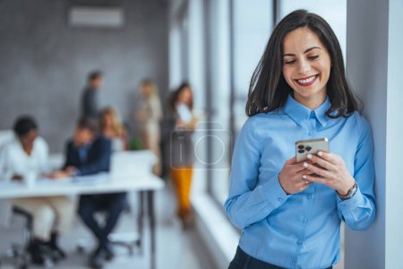 Photo for Beautiful business woman Texting while waiting the meeting. Shot of a young businesswoman using her phone at work. Smiling woman in casuals standing in office. Businesswoman with mobile phone in hand - Royalty Free Image
