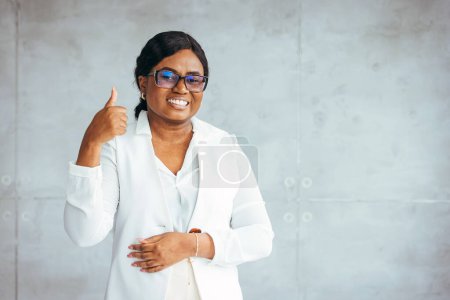 Téléchargez les photos : Portrait d'une jeune femme afro-américaine joyeuse portant un costume blanc souriant et regardant la caméra. Tournage d'une jeune femme d'affaires confiante travaillant dans un bureau moderne. - en image libre de droit