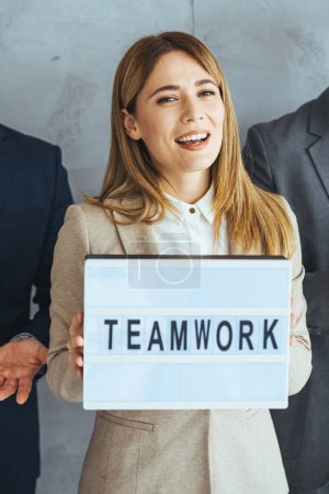 Téléchargez les photos : Portrait d'une équipe d'affaires multiethnique prospère regardant la caméra dans le bureau et le chef d'équipe tenant signe "travail d'équipe" dans les mains. Groupe d'entreprises multiethniques prospères - en image libre de droit