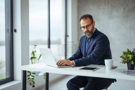 Photo for Cheerful adult man with glasses, taking notes on the conference call. Handsome young businessman looking thoughtful while working on his laptop in the office. Portrait of a handsome young businessman working in office - Royalty Free Image