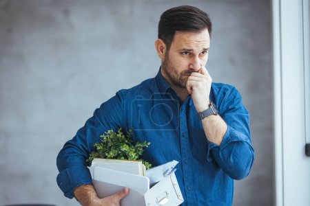 Photo for Business, Firing and Job Loss Concept - Sad Fired Male Office Worker With Box of His Personal Stuff. Fired male employee holding box of belongings in an office. Displeased mid adult businessman leaving the office - Royalty Free Image