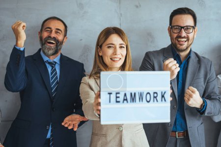 Photo for Portrait of multi ethnic successful business team looking at camera in the office and team leader holding sign ''teamwork'' in hands. Multi-ethnic group of business persons standing side by side - Royalty Free Image