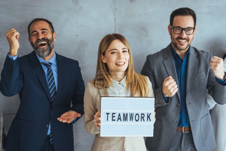 Photo for Portrait of multi ethnic successful business team looking at camera in the office and team leader holding sign ''teamwork'' in hands. Portrait of a group of businesspeople standing together in an office - Royalty Free Image