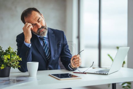 Photo for Frustrated mature  man massaging his head and keeping eyes closed while sitting at his working place in office. Exhausted businessman feeling pain after working on laptop. - Royalty Free Image