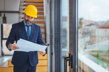 Photo for Young architect in suit with helmet and glasses holding blueprint in office - Royalty Free Image