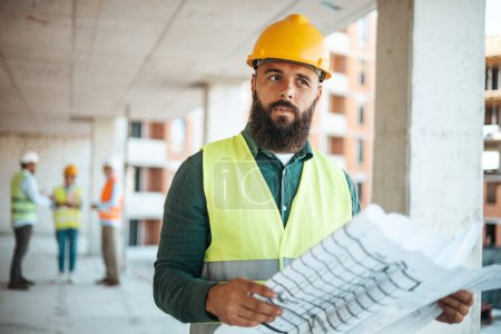 Photo for Bearded caucasian engineer with blueprint working at construction site with blurred colleagues on background - Royalty Free Image