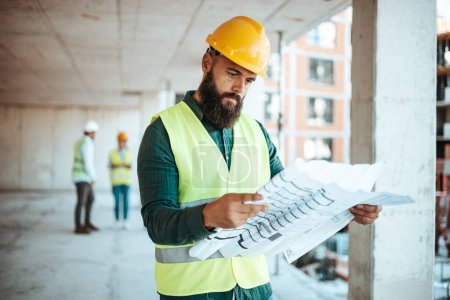 Téléchargez les photos : Ingénieur caucasien barbu avec un plan de travail sur le chantier de construction avec des collègues flous sur le fond - en image libre de droit