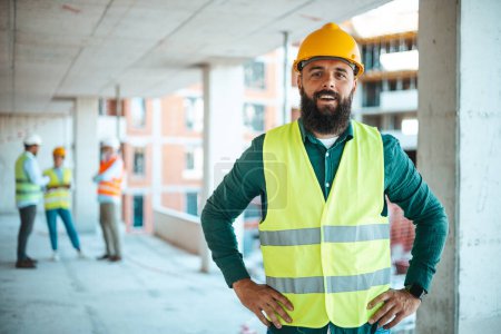 Photo for Portrait of bearded caucasian engineer with hands on hips working at construction site with blurred colleagues on background - Royalty Free Image