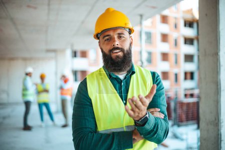 Foto de Retrato del ingeniero caucásico barbudo que trabaja en el sitio de construcción con colegas borrosos en el fondo - Imagen libre de derechos