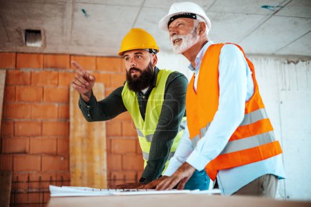 Téléchargez les photos : Ingénieurs caucasiens masculins ou architectes parlant pendant le travail avec le plan directeur sur le chantier de construction - en image libre de droit