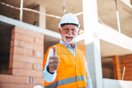 Téléchargez les photos : Portrait de l'ingénieur caucasien principal montrant pouce levé pendant le travail sur le chantier de construction avec des collègues flous sur le fond - en image libre de droit