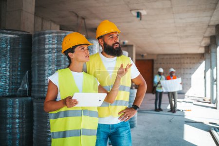 Photo for Young caucasian architects or engineers working with digital tablet at construction site with blurred colleagues on background - Royalty Free Image