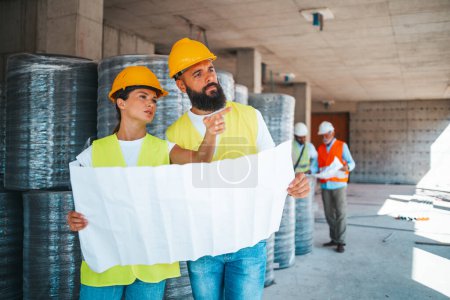 Photo for Young caucasian architects or engineers working with blueprint at construction site with blurred colleagues on background - Royalty Free Image