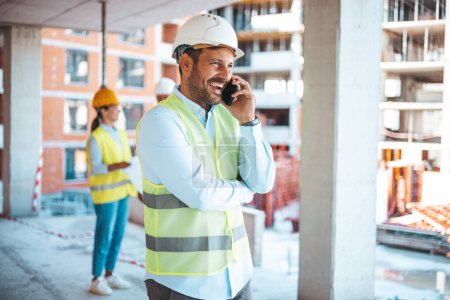 Photo for Smiling architect in suit and with helmet on head standing in building in construction process. Repair, building, construction, business and maintenance concept. Smiling man in helmet calling on smartphone  Building in construction process. - Royalty Free Image