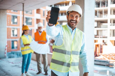 Photo for Engineer holding blueprints and showing a smartphone. Male construction engineer demonstrating his smartphone to camera with smile. Male engineer in hardhat looking at camera showing  smartphone - Royalty Free Image