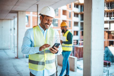 Shot of a young businessman using his smartphone while on a construction site. Portrait of young engineer in vest with white helmet standing on construction site, smiling and holding smartphone for worker, internet, social media. 