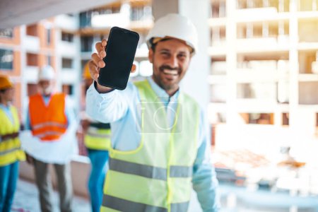 Photo for Engineer holding blueprints and showing a smartphone. Male construction engineer demonstrating his smartphone to camera with smile. Male engineer in hardhat looking at camera showing  smartphone - Royalty Free Image