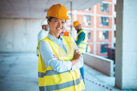 Photo for Portrait of woman engineer at building site looking at camera with copy space. Mature construction manager standing in yellow safety vest and yellow hardhat with crossed arms. Successful confident architect at construction site with team discussing i - Royalty Free Image