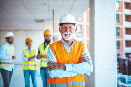 Photo for Portrait of a construction site worker in hard hat and safety vest. Senior architect at a construction site smiling at camera - Incidental people working at background. - Royalty Free Image