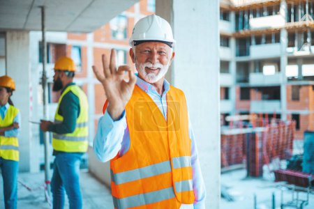 Photo for Young attractive architect standing at construction site and showing thumbs up. We're 100% safety compliant. Portrait of a senior man working showing an okay gesture at a construction site - Royalty Free Image