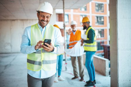 Photo for Smiling architect in suit and with helmet on head standing in building in construction process next to window and typing message on smart phone. Building in construction process. - Royalty Free Image