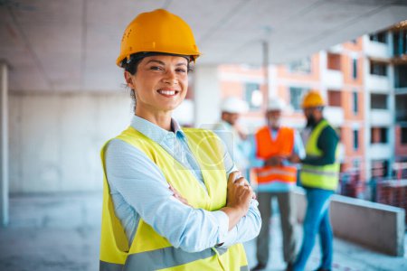 Photo for Portrait of woman engineer at building site looking at camera with copy space. Mature construction manager standing in yellow safety vest and yellow hardhat with crossed arms. Successful confident architect at construction site with team discussing i - Royalty Free Image