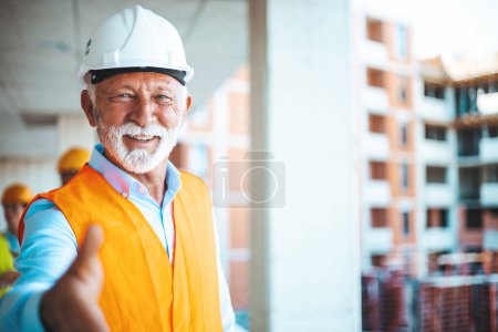 Foto de Retrato de un gerente de construcción. Él ofrece su mano para la conclusión de un nuevo negocio, detrás de él los trabajadores de la construcción viendo planes con el arquitecto. - Imagen libre de derechos