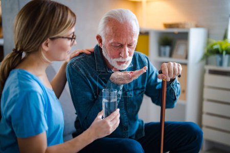 Photo for A home healthcare worker helping a senior man in his 70s take his prescription medicine. The patient is in the kitchen, holding a cup of water, looking down as the nurse puts the pills in his hand. - Royalty Free Image