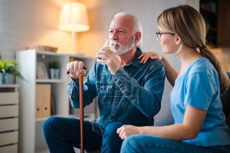 Photo for A young nurse sits at a table in the living room with a senior man and provides him with medical assistance. Female caregiver giving the medicine to her older male patient - Royalty Free Image