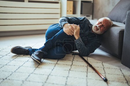Téléchargez les photos : Ancien homme âgé couché sur le sol après être tombé avec un bâton de marche en bois à côté du canapé sur le tapis dans le salon à la maison. Vieil homme souffrant de douleur et luttant pour se lever après être tombé à la maison - en image libre de droit