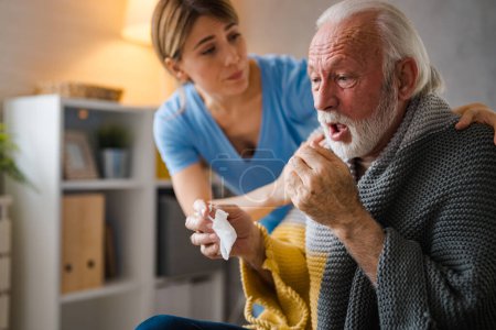 Photo for Cold exhausted senior man with flu wrapped in a warm blanket blowing his nose with a tissue in the livingroom. Nurse take care of him. Geriatrician helping lonely elderly sick man. - Royalty Free Image