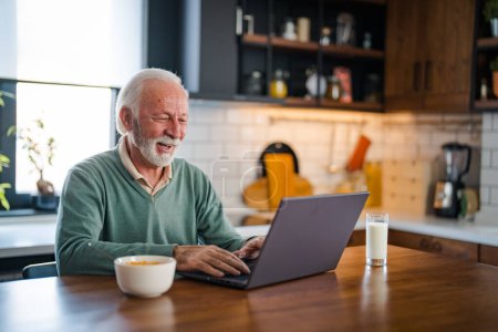 Photo for Senior man working at laptop at home. Retired Man On Phone At Home In Kitchen Using Laptop Celebrating Good News. Senior adult in home kitchen with tea cup and laptop. - Royalty Free Image